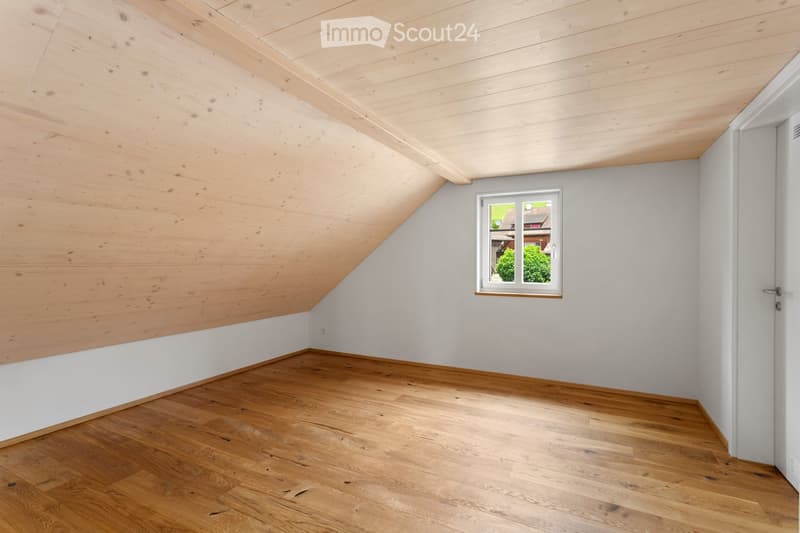 Empty attic room with wooden floors, white walls, and a skylight window. The ceiling has exposed wooden beams, and the walls are painted in a light gray color with a wooden panel accent along the lower portion.