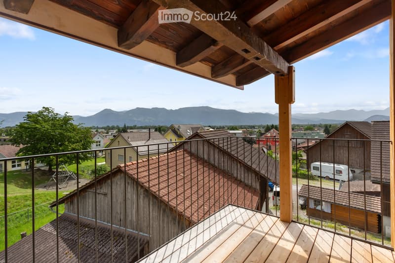 Balcony with wooden decking and metal railing, overlooking a town with houses, trees, and distant mountains.