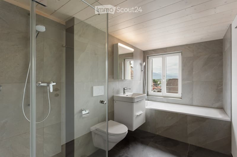 Modern bathroom with grey tiles, a glass-enclosed shower, a white sink, a toilet, and a large window offering a view.