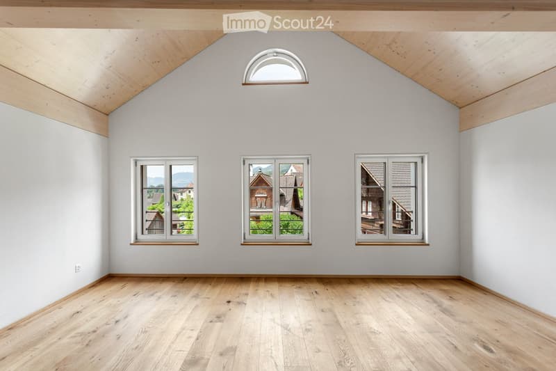 Empty room with wooden flooring, white walls, and multiple windows offering views of the outside. The ceiling features exposed wooden beams and a skylight.