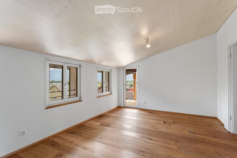 Empty room with wooden flooring, white walls, and large windows offering a view of the landscape. The room features a door leading to a balcony or terrace area.
