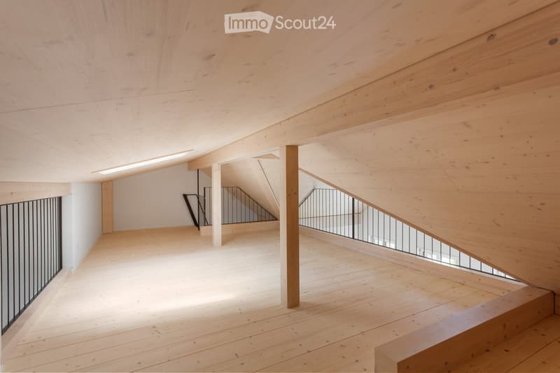 Open, airy hallway with wooden floors and ceilings, featuring a staircase with metal railings and wooden balusters, leading to an upper level.