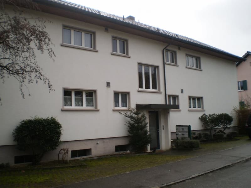 Two-story residential building with white exterior walls, multiple windows, and a small entrance area with a mailbox. The building has a tiled roof and is surrounded by small shrubs and trees.