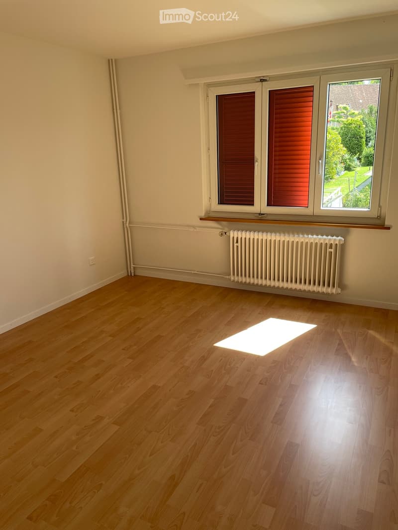 Empty room with light wooden flooring, white walls, and a radiator under a large window with red shutters. The room has a simple, clean design with no furniture.