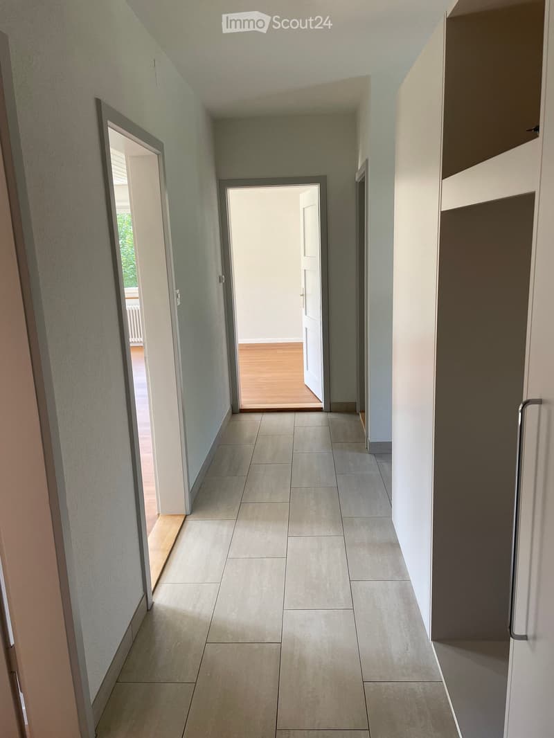 Clean, modern hallway with tiled flooring and light-colored walls. Features multiple doors leading to different rooms, including a visible wooden floor room at the end.