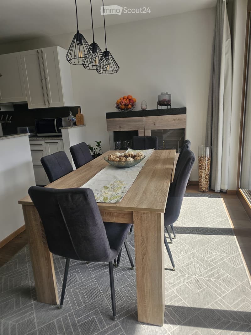 Modern dining area with a wooden dining table, six dark grey chairs, a decorative bowl with decorative items, a fireplace, and pendant lights. The area is connected to a kitchen with white cabinets and a microwave.