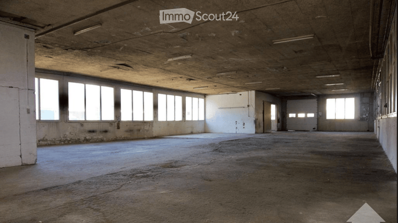 Empty industrial-style room with concrete floors and walls, large windows, and exposed ceiling beams. No furniture or equipment present.