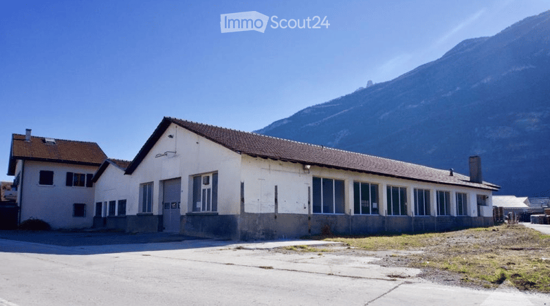 A two-story building with a flat roof and large windows, situated in a rural area with a mountain in the background.