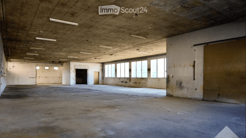 Empty industrial-style room with concrete floors and walls, large windows, and exposed ceiling with fluorescent lighting.