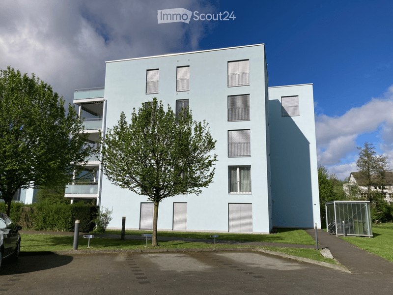 Modern, light blue apartment building with multiple floors, balconies, and large windows. The building is surrounded by greenery and has a paved parking area.