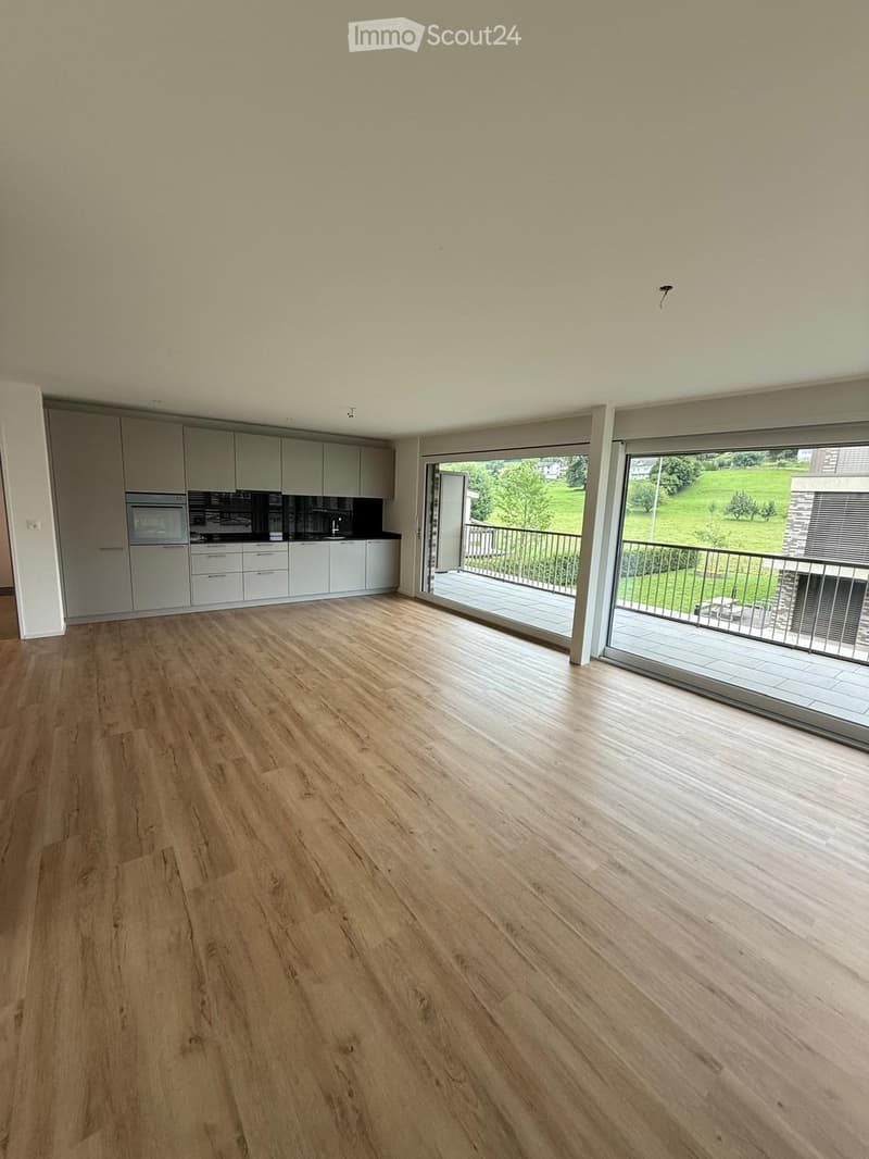 Empty room with light wooden flooring, white walls, and large sliding glass doors leading to a balcony with a view of a green area.