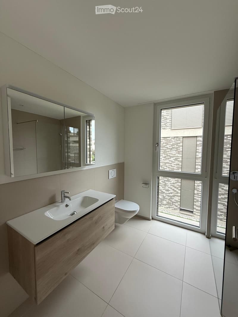 Modern bathroom with a double sink, wooden cabinet, mirror, toilet, and large window with blinds.