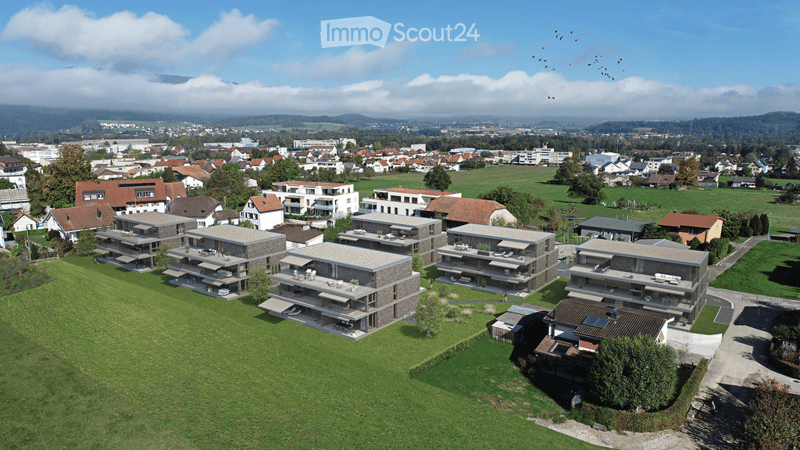 Aerial view of a residential area with modern apartment buildings, green lawns, and surrounding town with houses and fields.