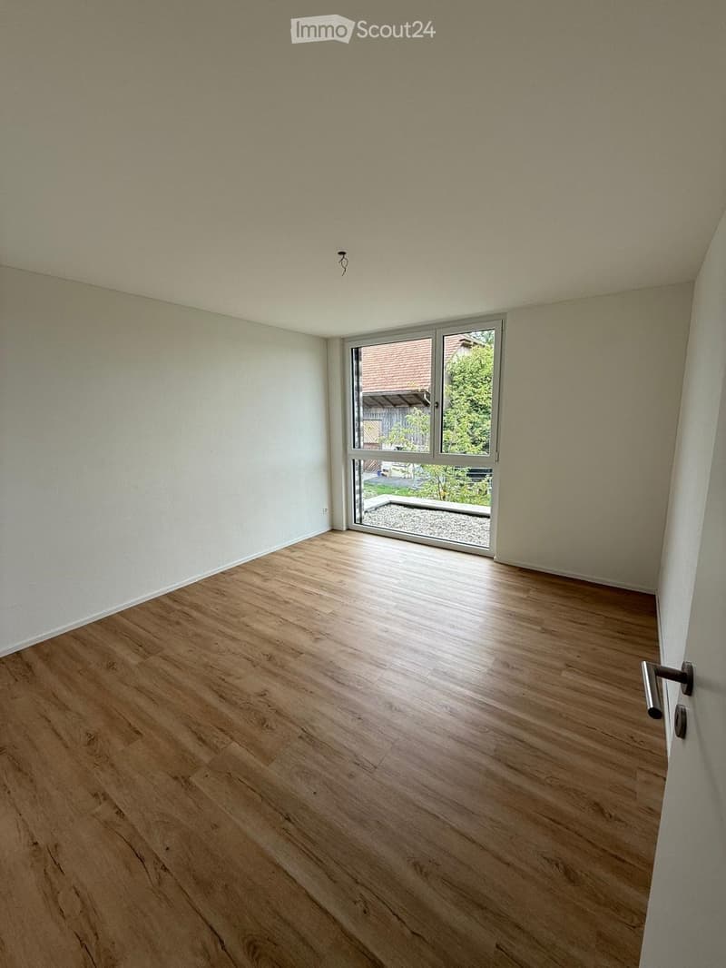 Empty room with wooden flooring and white walls, featuring a large window with a view of a garden area.