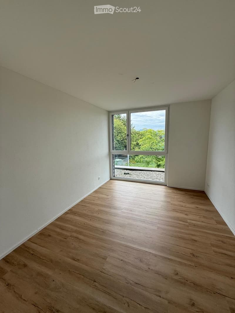Empty room with wooden flooring, white walls, and large window offering a view of greenery outside.