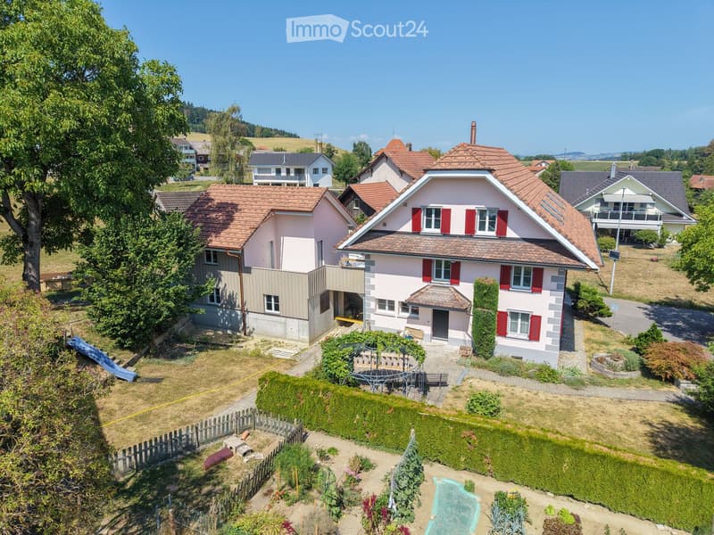 A two-story house with a red-tiled roof and white walls, surrounded by a well-maintained garden and other houses in the background.