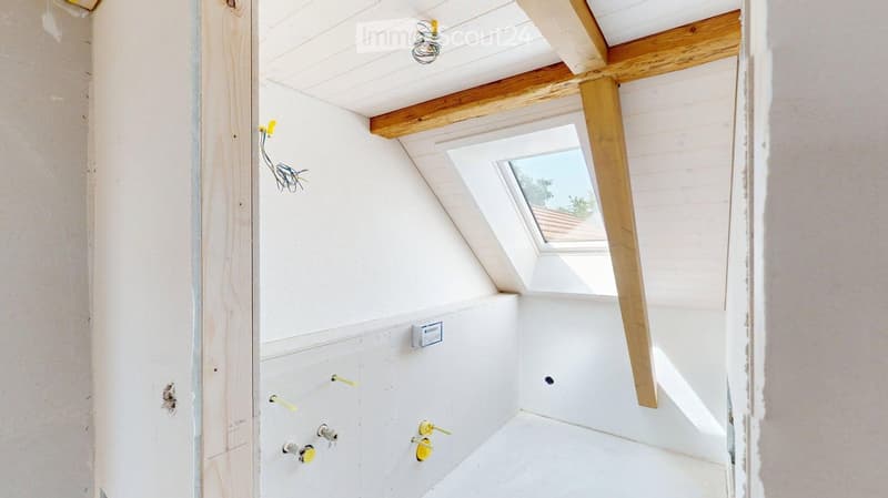 Empty attic room with white walls, wooden beams, and a skylight window. Exposed wiring and unfinished surfaces visible.