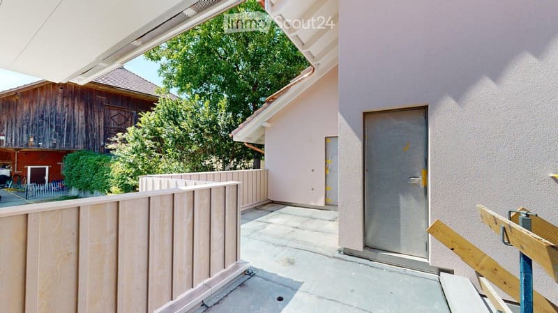 Balcony with wooden railing, concrete floor, and a view of a wooden shed and greenery.