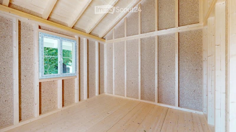 Empty room with exposed wooden beams, wooden floor, and a single window with a view of greenery outside.