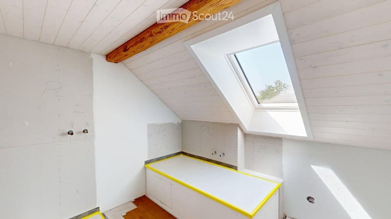 Empty room with white walls, wooden ceiling beams, skylight window, and a bathtub base with yellow tape marking.