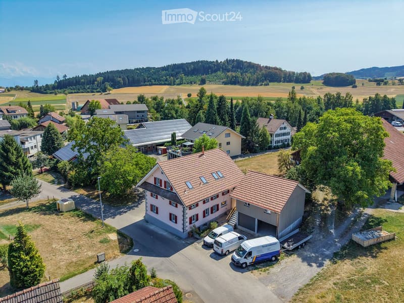 Aerial view of a two-story house with a red-tiled roof, surrounded by trees and other houses in a rural area. A garage is attached to the house, and several vans are parked in the driveway.