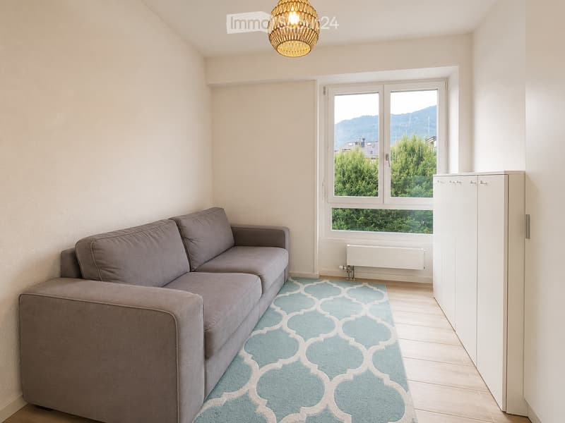 Modern living room with a grey sectional sofa, a blue patterned rug, and a white cabinet. Large windows offer a scenic mountain view.