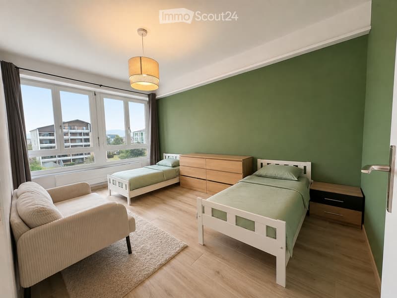 Bright bedroom with two single beds, a sofa, a dresser, and a nightstand. Green accent wall, large windows with curtains, and a hanging light fixture.