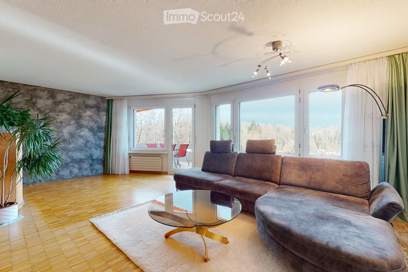 Modern living room with large windows, wooden flooring, a grey sectional sofa, a glass coffee table, and a floor lamp. A potted plant is visible on the left side.