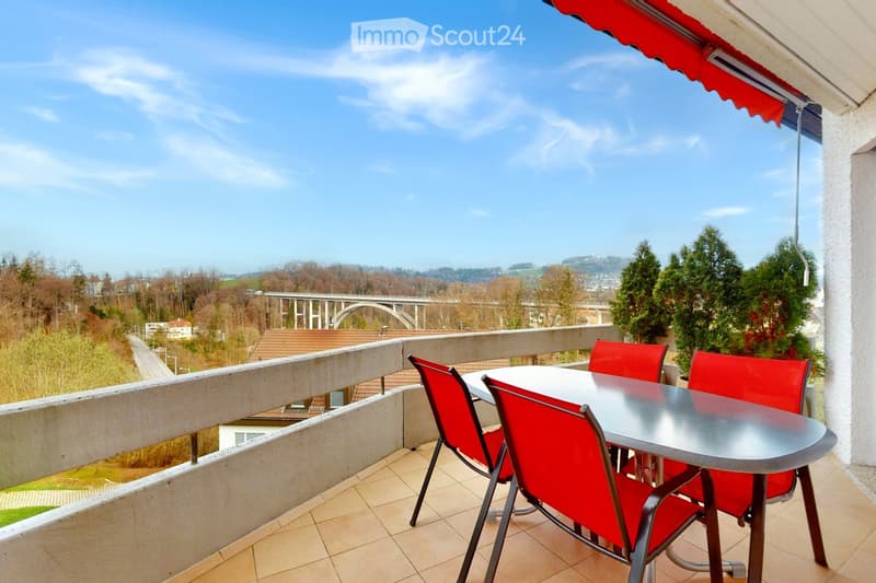Balcony with a table and four red chairs, overlooking a scenic landscape with a bridge and trees.