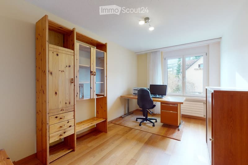 A well-lit study or home office with wooden flooring, a large wooden cabinet with drawers and shelves, a desk with a chair, a laptop, a printer, and a radiator under the window.