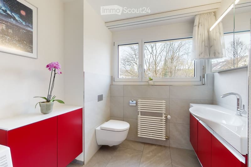 Modern bathroom with a white sink, toilet, radiator, red cabinets, and a window with a view of trees. A flower pot with purple orchids is placed on the counter.