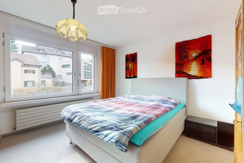Bedroom with a large window, a bed with a colorful blanket, a nightstand, and two paintings on the wall.