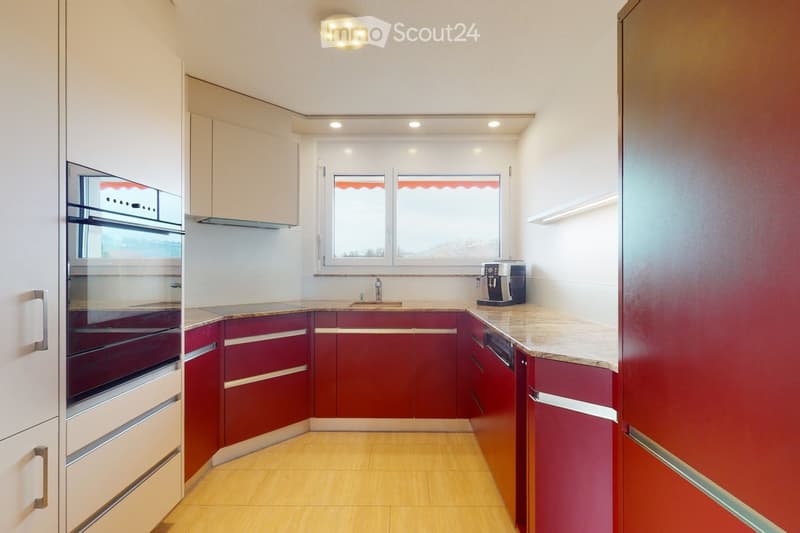 Modern kitchen with red cabinets, white walls, wooden flooring, and a large window. Features an oven, sink, faucet, and a coffee maker on the counter.