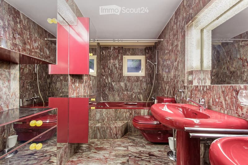 Bathroom with red fixtures, marble walls and floor, two sinks, two toilets, and a mirror.