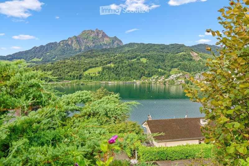 Scenic view of a lake surrounded by lush green hills and mountains, with a small house visible in the foreground.