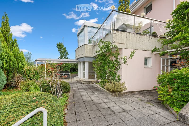 A modern house with a curved design and a glass balcony, surrounded by lush greenery and a paved pathway leading to the entrance.