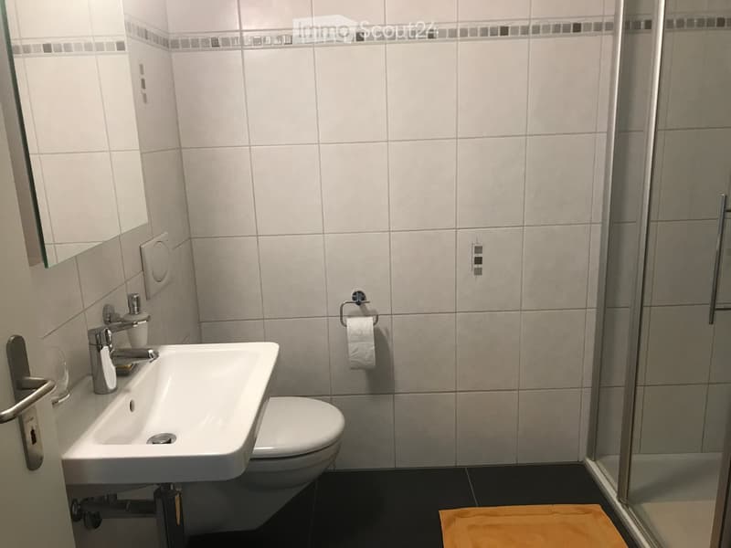 Modern bathroom with white tiles, a sink with a chrome faucet, a toilet, a glass-enclosed shower, and a towel on the floor.