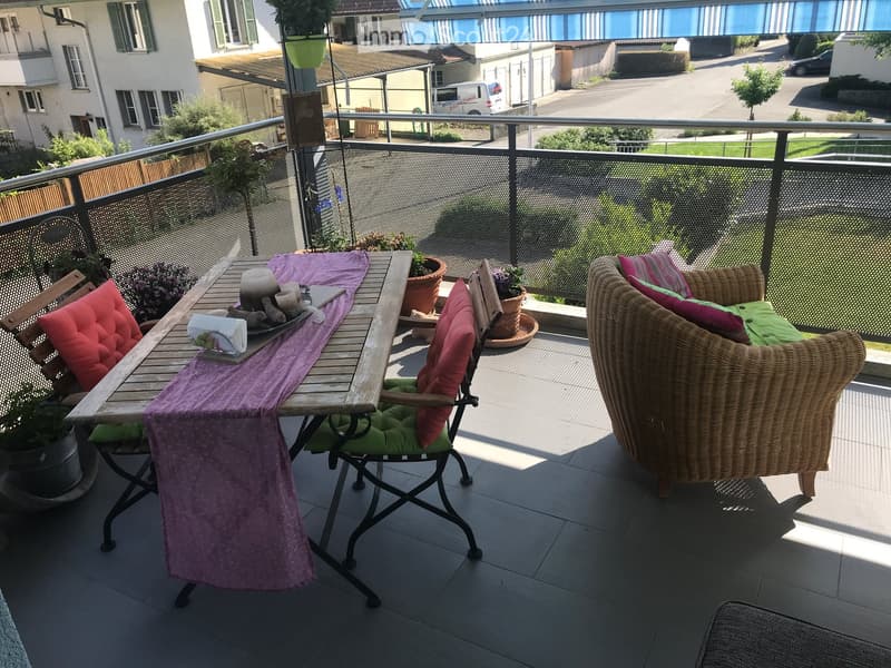 Balcony with a wooden table, two chairs, a wicker chair, cushions, and potted plants.