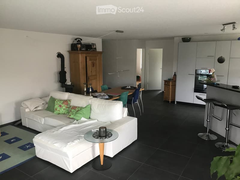 Modern living room with a white sectional sofa, green throw pillows, a glass coffee table, and a small round table. The room features a dining area with a wooden table and chairs, a kitchen area with white cabinets and a black countertop, and a small bar