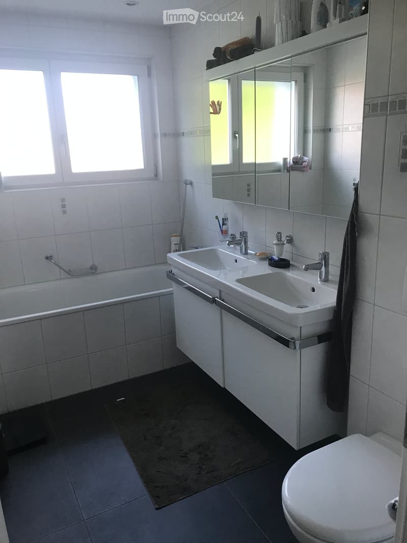 Modern bathroom with white tiles, double sink, bathtub, toilet, large mirror, and towel on the side.
