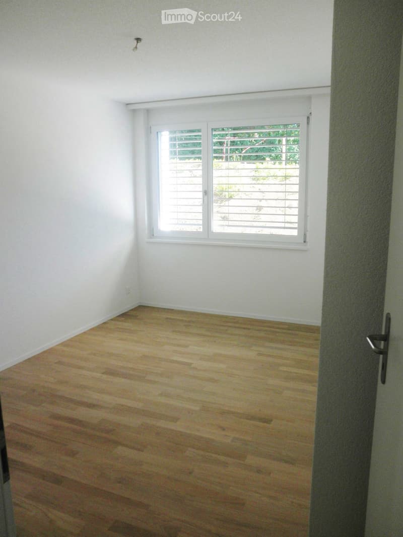Empty room with light wooden flooring and white walls, featuring a large window with blinds and a partially visible door frame.