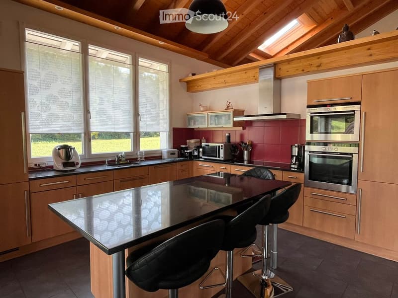 Modern kitchen with wooden cabinets, stainless steel appliances, a central island with bar stools, and large windows with blinds.