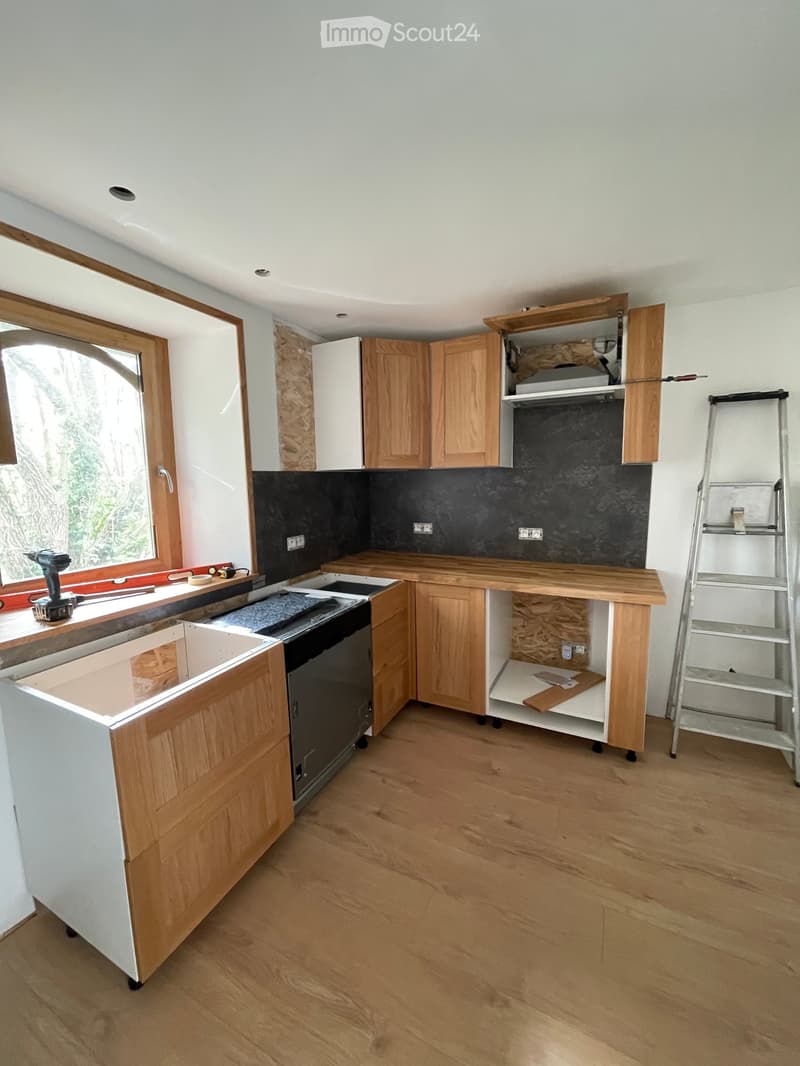 Kitchen with wooden cabinets, a sink, and a drill on the windowsill. The floor is wooden, and there is a ladder on the right side.
