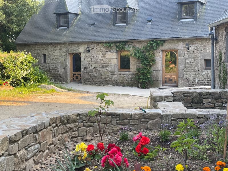 Stone house with a slate roof, multiple windows, and wooden doors, surrounded by a garden with colorful flowers and a stone pathway.