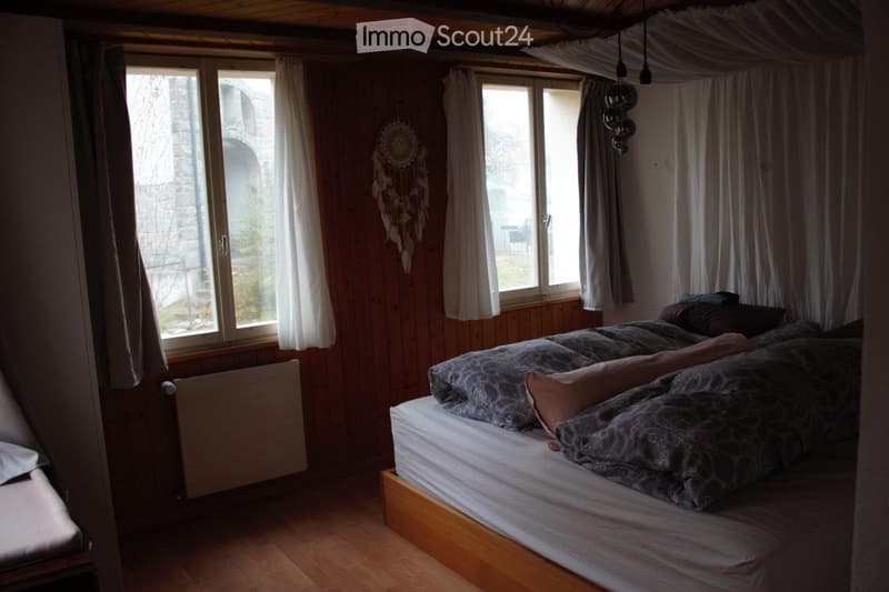 Cozy bedroom with wooden walls and floor, large windows with curtains, a double bed with patterned bedding, a dreamcatcher on the wall, and a radiator below the window.