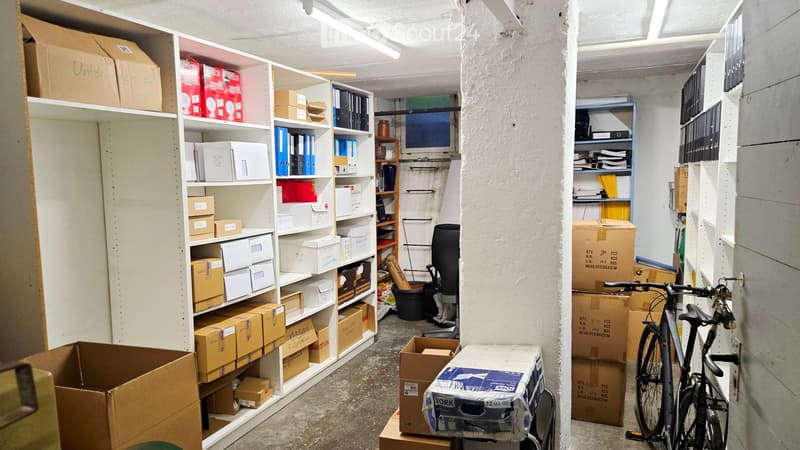 Storage room with white shelving units filled with cardboard boxes, storage bins, and office supplies. A bicycle is leaning against the wall, and an office chair is positioned near a small window.