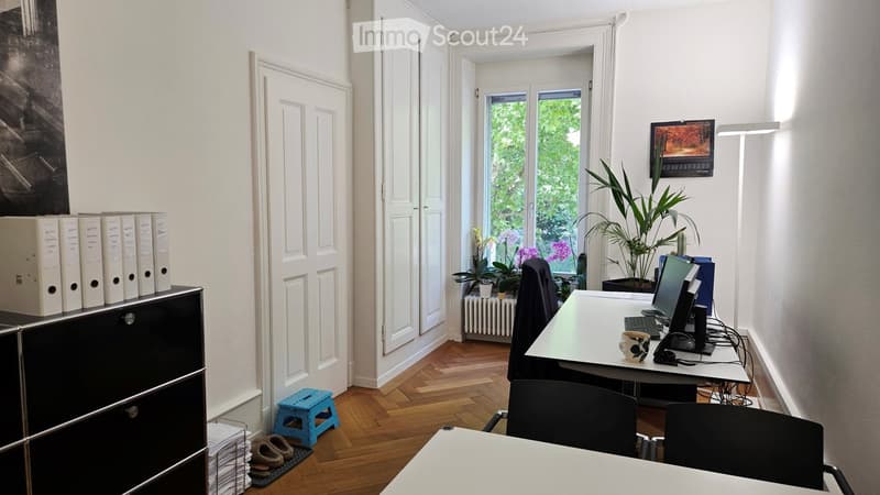 A well-lit office space with a wooden floor, white walls, and large windows. The room features a desk with a computer, keyboard, and a cup, along with a chair, a small stool, and a potted plant. A cabinet with files and a screen is also visible.