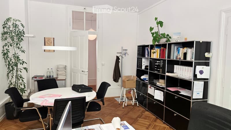 Office space with a round table, chairs, laptop, bookshelf, potted plants, and various office supplies.