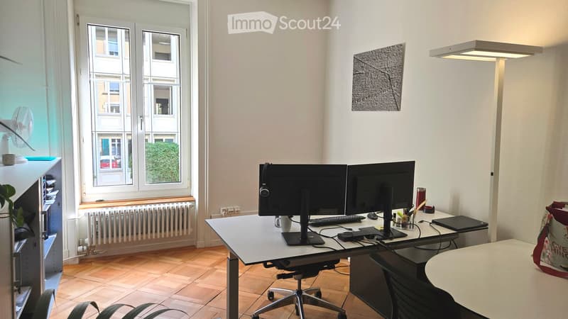 A modern, well-lit office space with a large wooden desk, dual monitors, a laptop, keyboard, mouse, and a comfortable chair. The room features large windows with a view of the street, a fan, a potted plant, and a cabinet with office supplies.