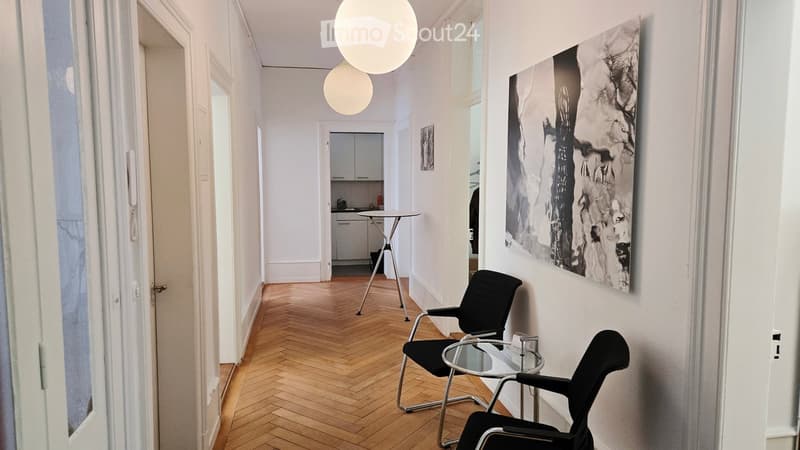 A modern, minimalist hallway with herringbone wooden flooring, white walls, and large windows. Features include a black chair, a small glass table, a stool, a cabinet, and an art piece on the wall.